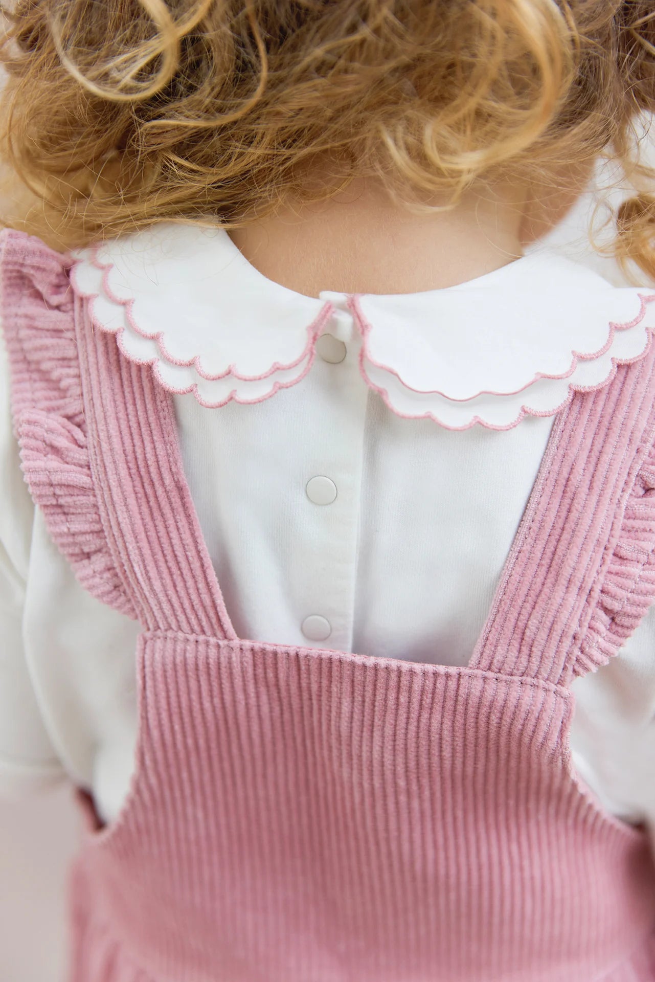 Close-up of a young child from behind, showing curly hair and a white shirt with scalloped collar, wearing Tartine et Chocolat's Baby Girl's Velvet Overalls in eglantine pink with ruffle straps.