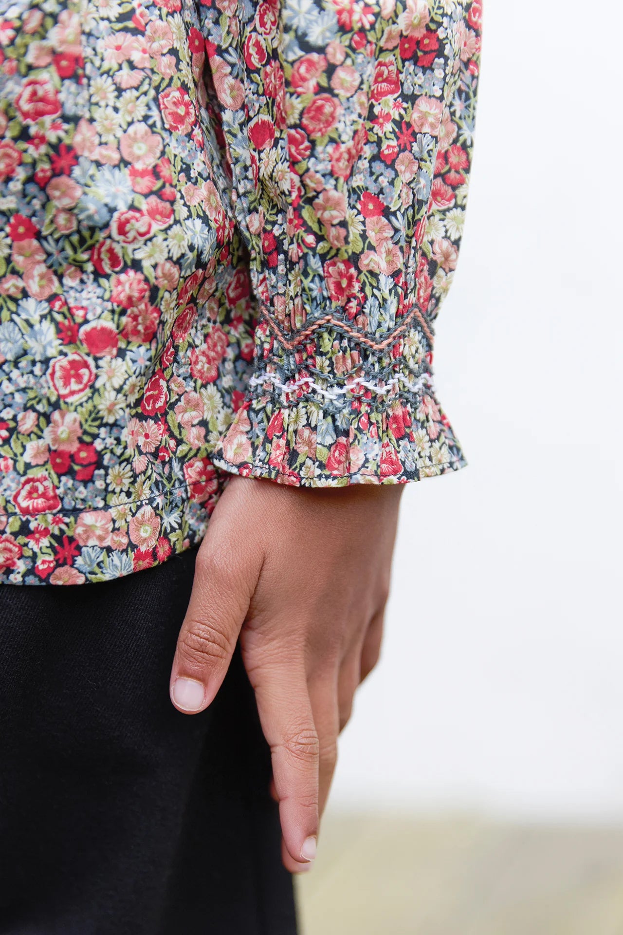 A close-up of a girl’s hand by her side, wearing the Tartine et Chocolat Girls' Liberty Smocked Floral Tunic with ruffled cuffs and black pants.
