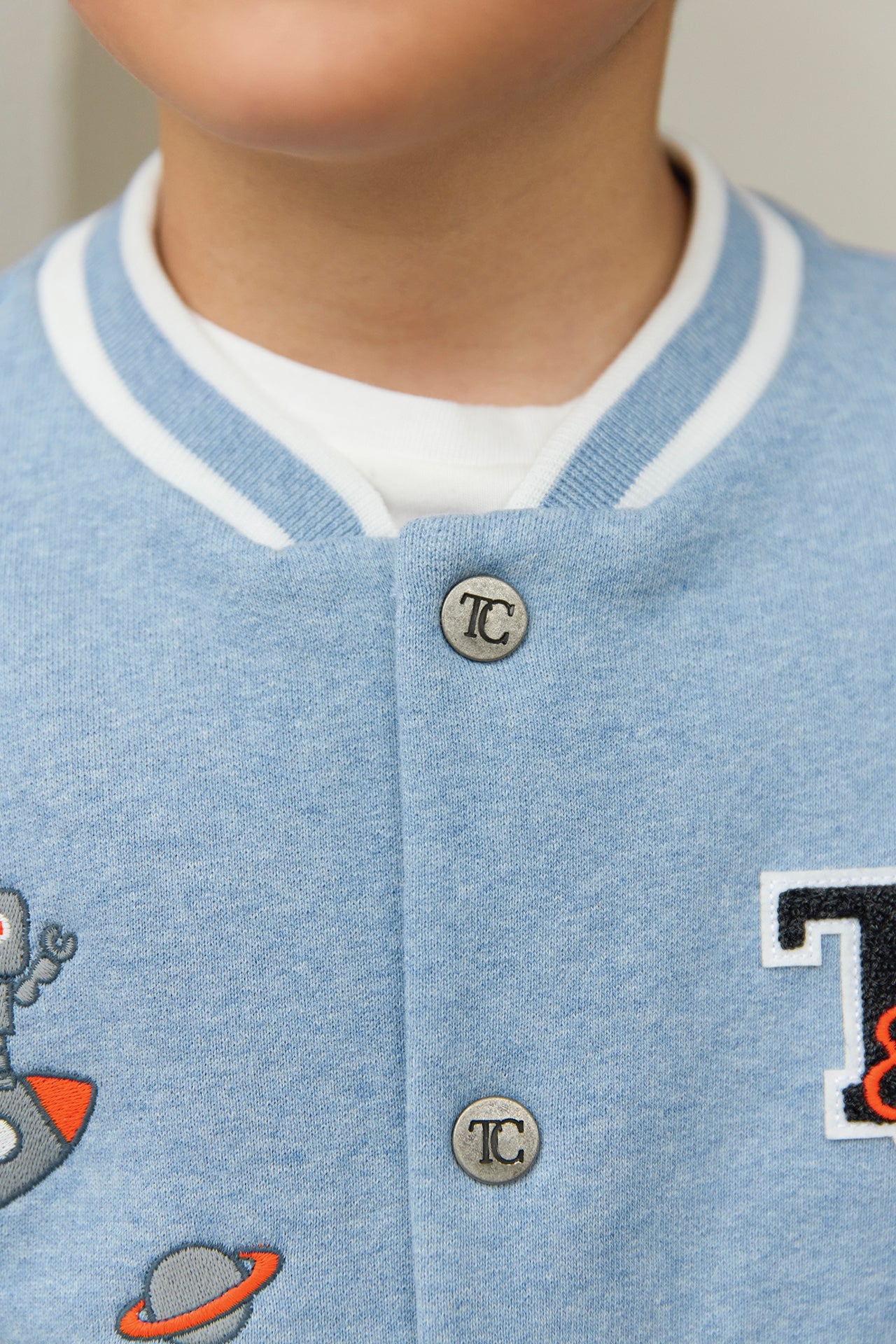 Close-up of a child in Tartine et Chocolat’s Baby Boys' and Boys' Easy Bomber Cardigan, light blue with white trim, metal snap buttons, and embroidered robot, planet, and "T" designs.