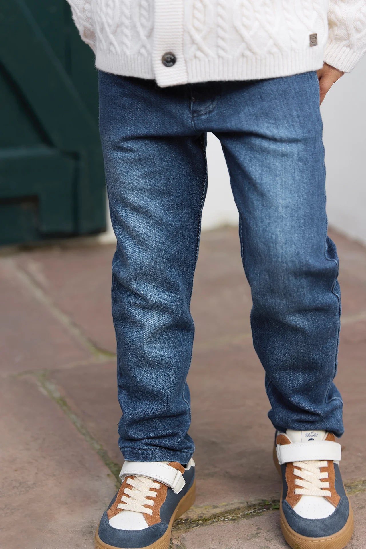 A child in Tartine et Chocolat Baby Boys' and Boys' Cotton Jeans, a white knit sweater, and blue-brown sneakers stands on stone pavement—perfect for everyday wear.