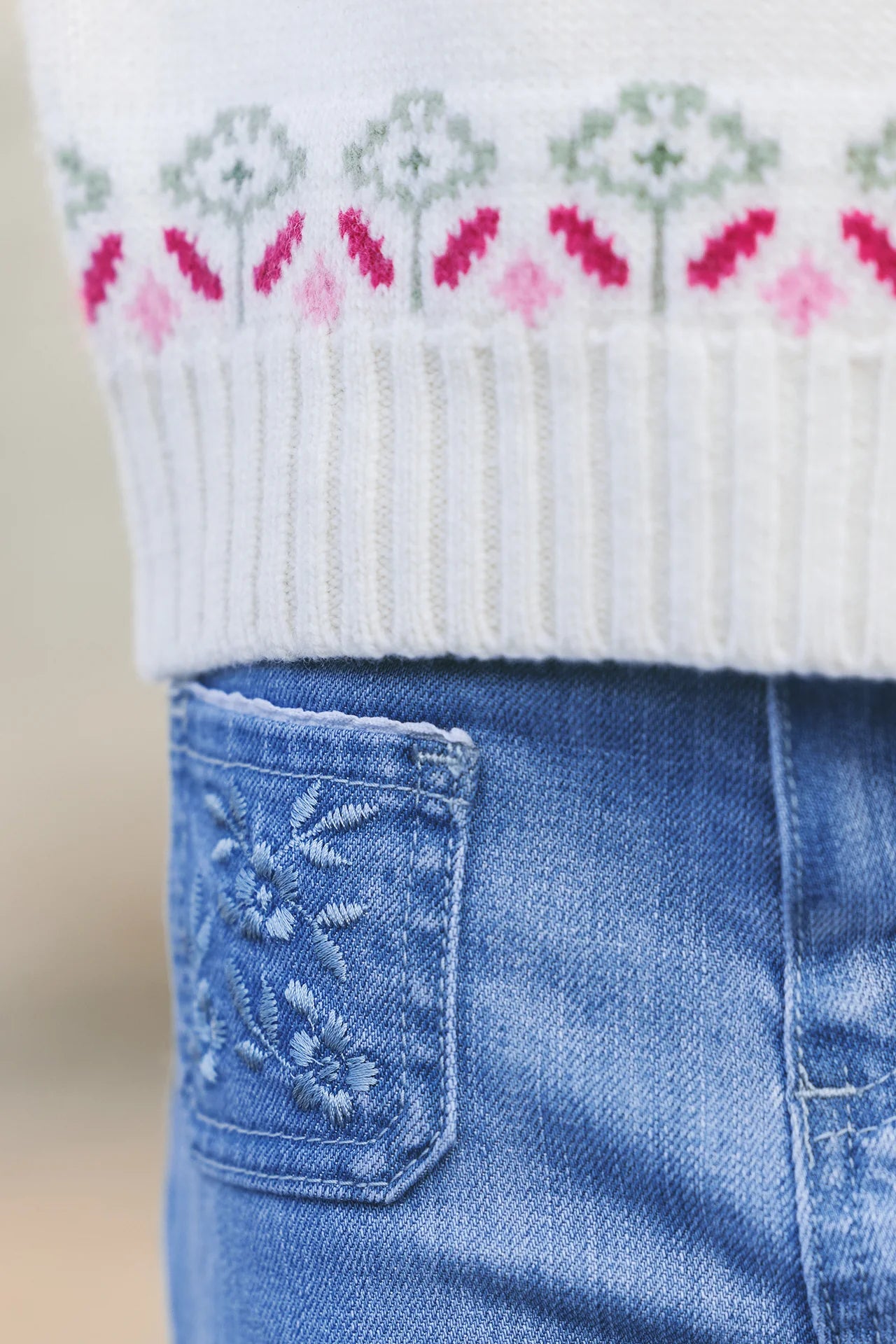Close-up of Tartine et Chocolat Girls' Blue Flared Embroidery Jeans with floral detailing on the pocket, paired with a white knit sweater featuring a pink and green pattern.