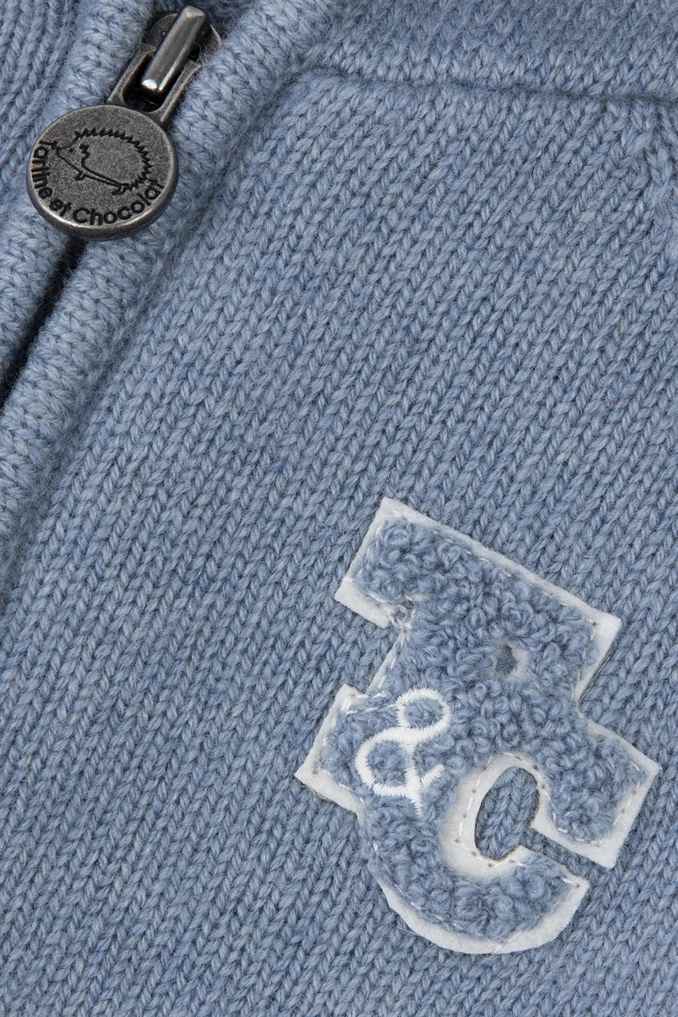 Close-up of the Tartine et Chocolat Baby Boys' and Boys' TC Signature Knit Zip Jacket, featuring a blue-grey knit, zipper with round metallic pull, and textured "C" and "8" appliqué—perfect for stylish layering.