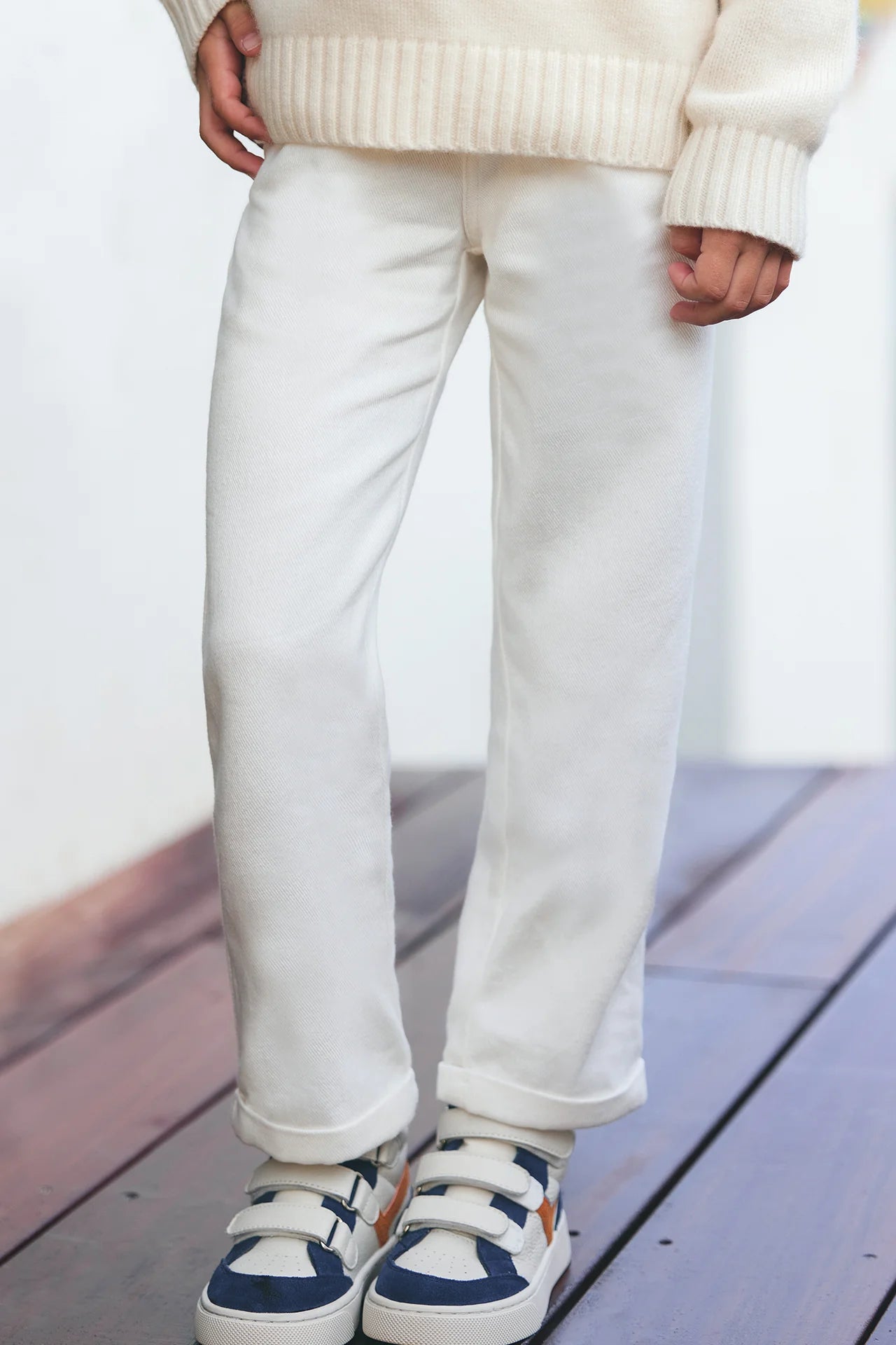 A junior girl wearing Tartine et Chocolat’s Girls' Drawstring Bow Trousers stands on a wooden floor, paired with a cream knit sweater and blue-and-white sneakers.