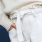 Close-up of a person wearing an off-white textured sweater and Tartine et Chocolat Girls' Drawstring Bow Trousers in ivory, featuring a fabric belt tied in a bow, with hand in pocket.