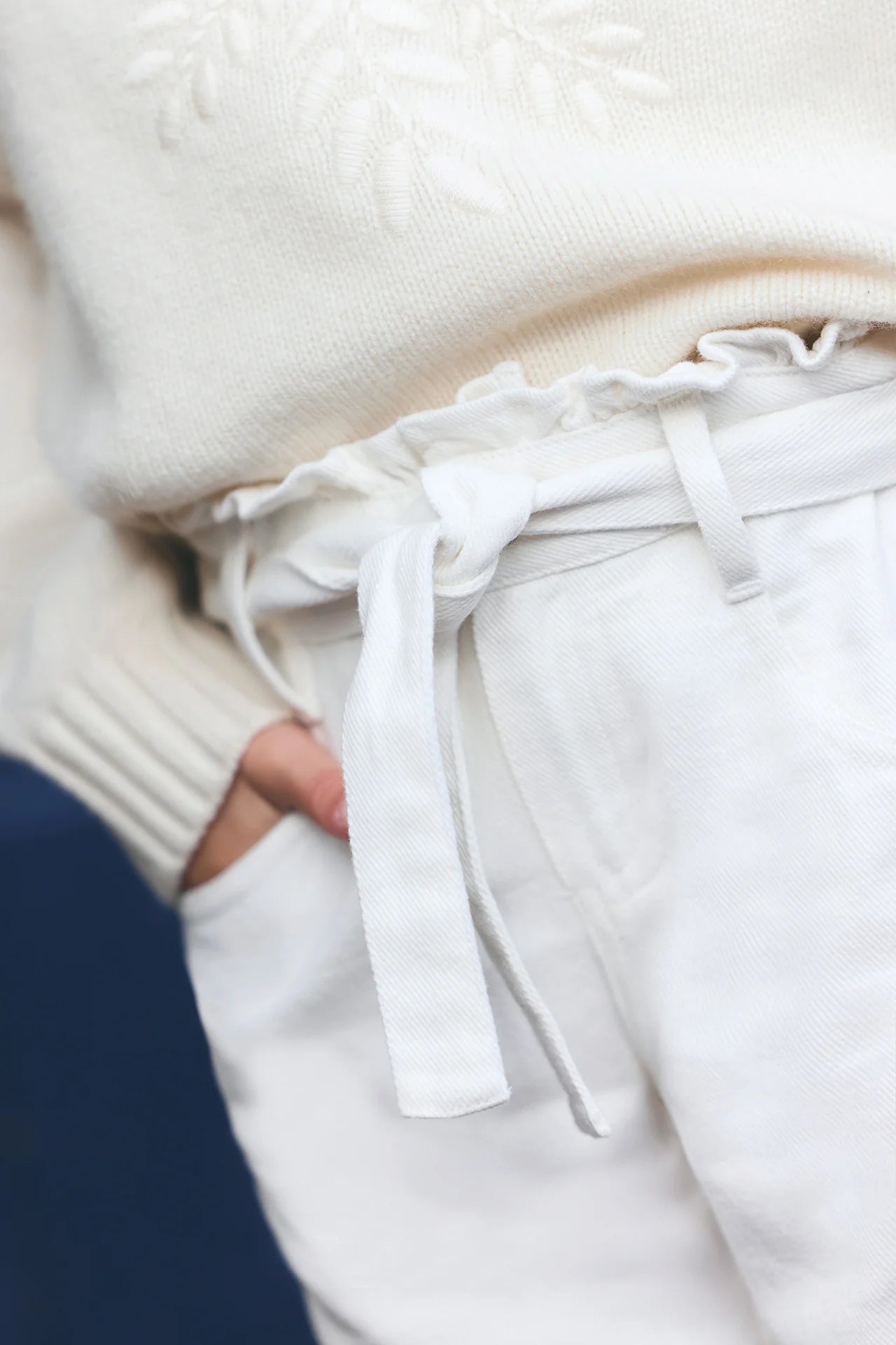 Close-up of a person wearing an off-white textured sweater and Tartine et Chocolat Girls' Drawstring Bow Trousers in ivory, featuring a fabric belt tied in a bow, with hand in pocket.