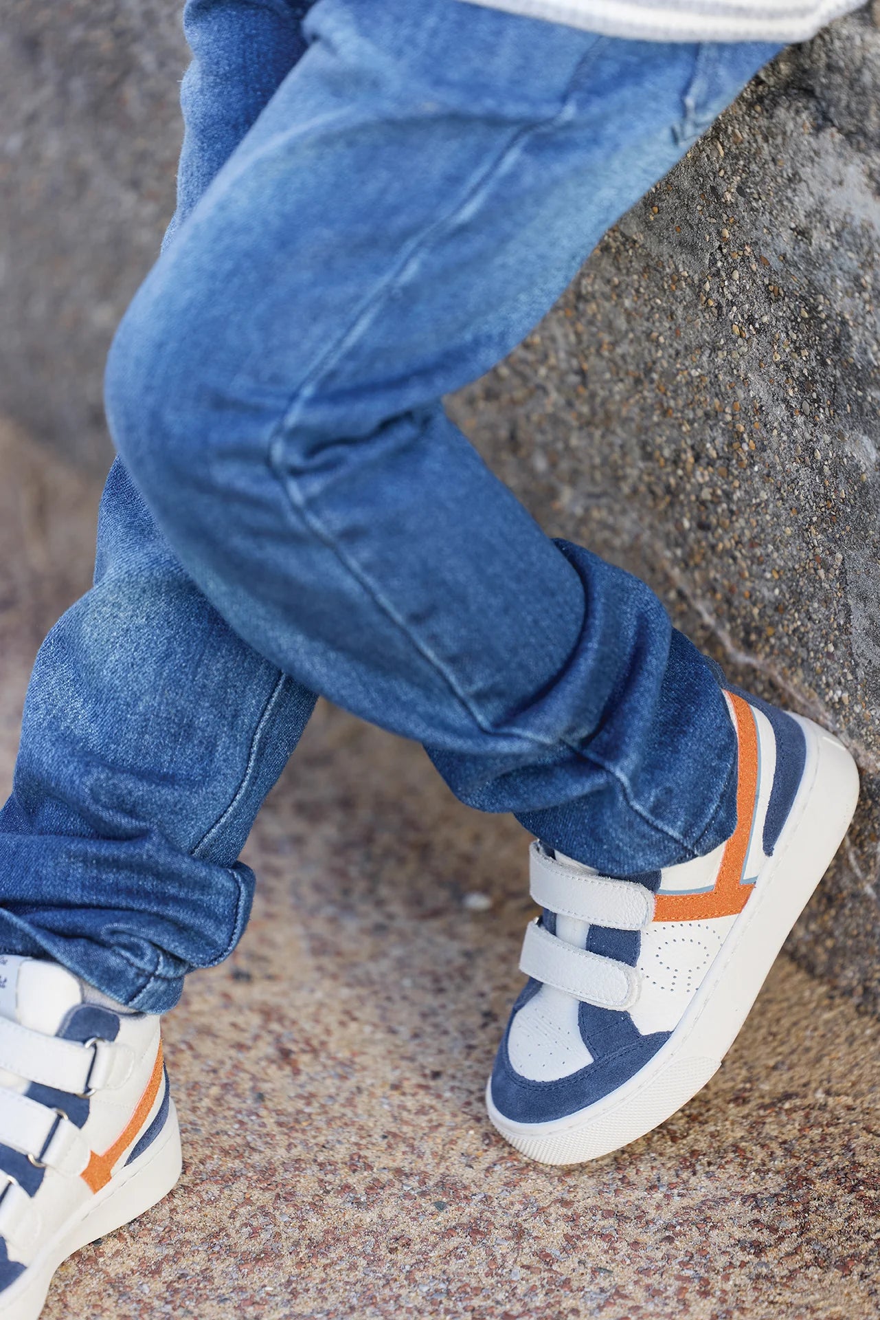 A child in Tartine et Chocolat Baby Boys' and Boys' Cotton Jeans sits cross-legged on a stone surface, wearing blue jeans and white sneakers with orange accents.