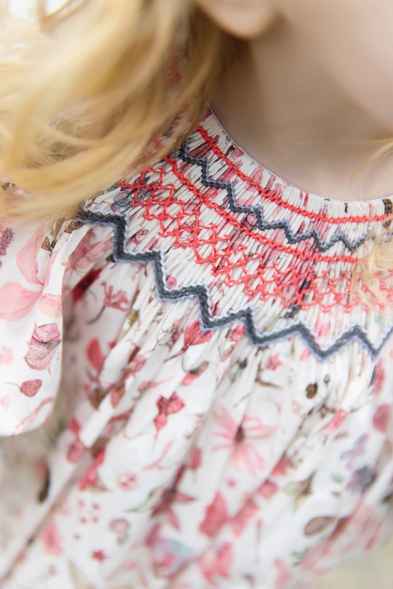 Close-up of a child in a Tartine et Chocolat Baby Girls' and Girls' Smocked Collar Floral Dress, showing detailed smocking with red and blue embroidery on the collar; part of her blonde hair is also visible.