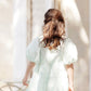 A young girl with brown hair wears a Tartine et Chocolat Baby Girls' and Girls' Puff Sleeve Dress and holds a woven bag while standing outdoors, facing away from the camera.
