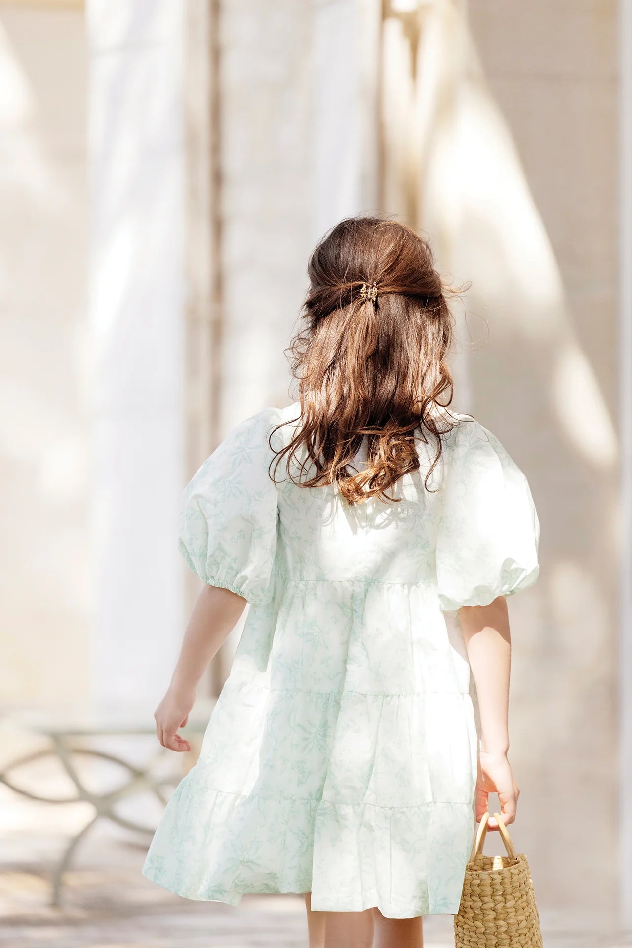A young girl with brown hair wears a Tartine et Chocolat Baby Girls' and Girls' Puff Sleeve Dress and holds a woven bag while standing outdoors, facing away from the camera.