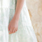 A girl wears the Tartine et Chocolat Baby Girls' and Girls' Puff Sleeve Dress in white and light green, standing with one arm relaxed against a beige textured background.