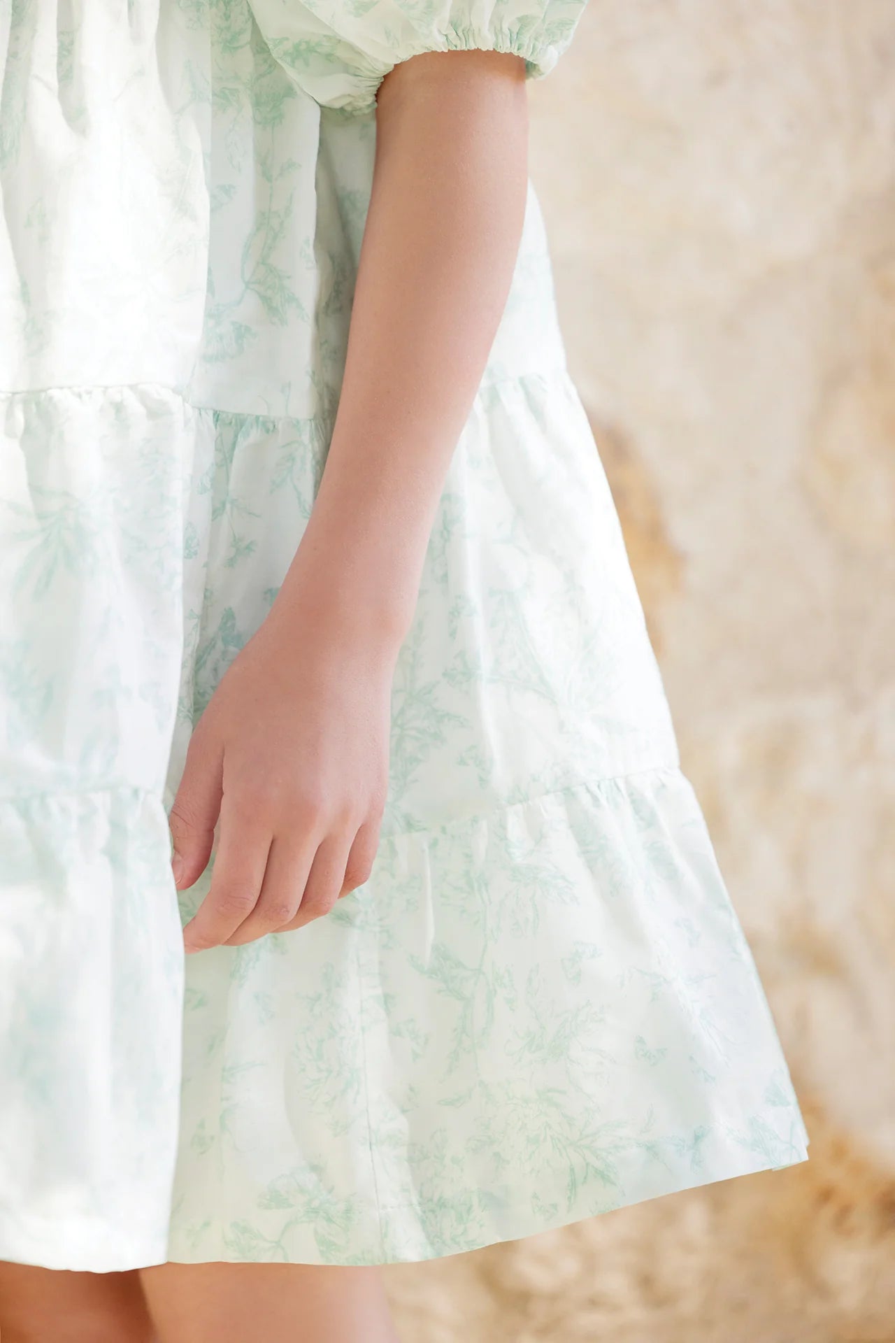 A girl wears the Tartine et Chocolat Baby Girls' and Girls' Puff Sleeve Dress in white and light green, standing with one arm relaxed against a beige textured background.