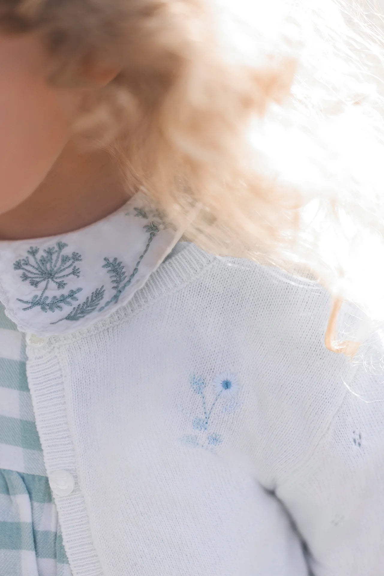 Close-up of a child in a green gingham dress and collared shirt, wearing the Tartine et Chocolat Baby Girls' Emeraude Embrodery Floral Cardigan, which beautifully complements her curly blond hair.
