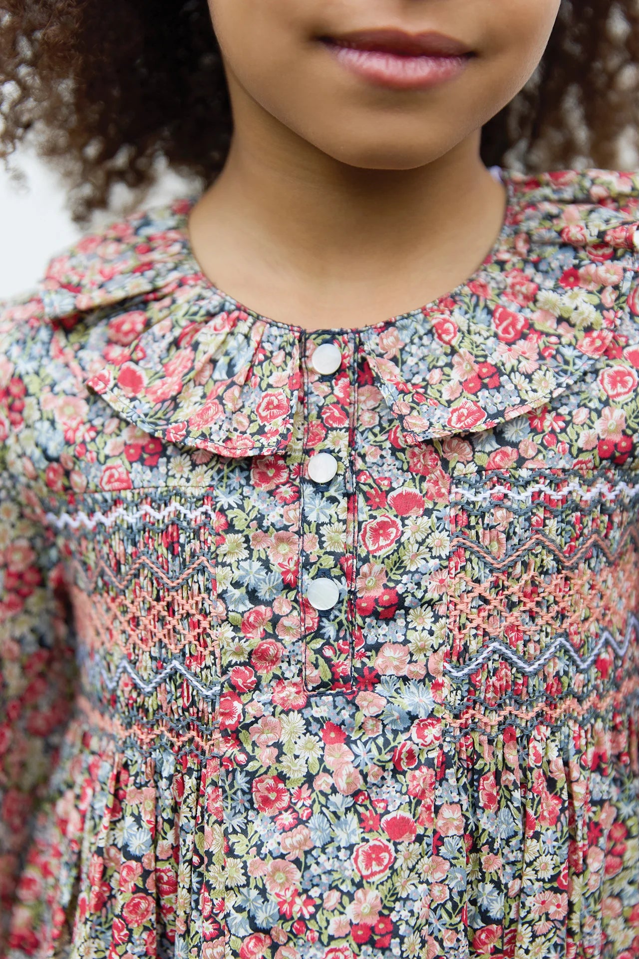 A girl models the Tartine et Chocolat Girls' Liberty Smocked Ruffle Sleeved Floral Dress, featuring decorative buttons and a charming floral print—elegantly shown from shoulders to chin.