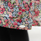 A close-up of the Tartine et Chocolat Girls' Liberty Smocked Ruffle Sleeved Floral Dress with a colorful floral print and white button detail, styled over black pants.