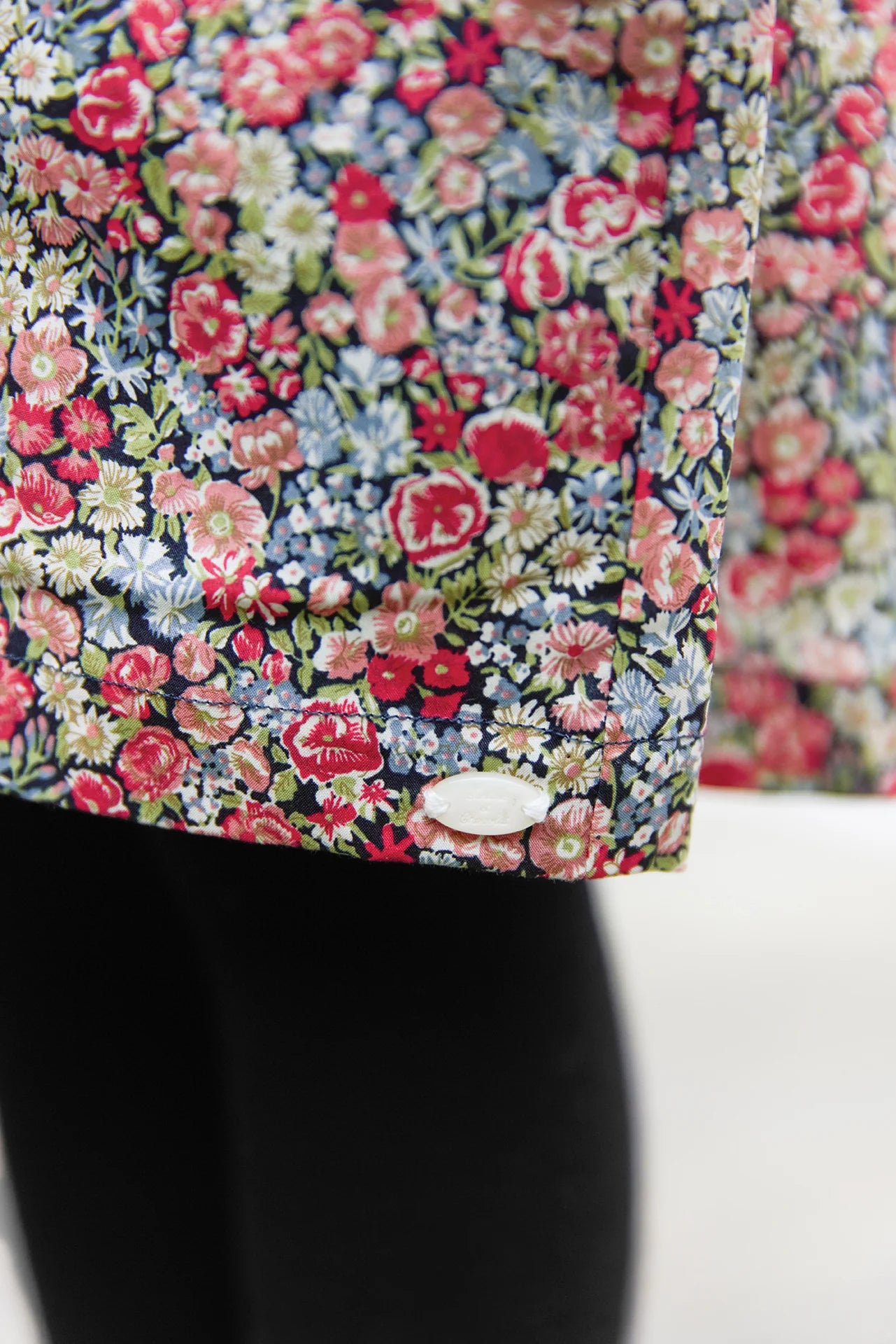 A close-up of the Tartine et Chocolat Girls' Liberty Smocked Ruffle Sleeved Floral Dress with a colorful floral print and white button detail, styled over black pants.