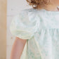 A young child is shown from the shoulders down, wearing the Tartine et Chocolat Baby Girls' and Girls' Puff Sleeve Dress with a subtle floral pattern; part of her face is out of frame.
