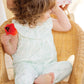 A young child with wavy brown hair sits in a wicker chair, wearing the Tartine et Chocolat Baby Girls' Sleeveless Jumpsuit and holding a red flower, looking to the side. An adult stands partially visible in the background.