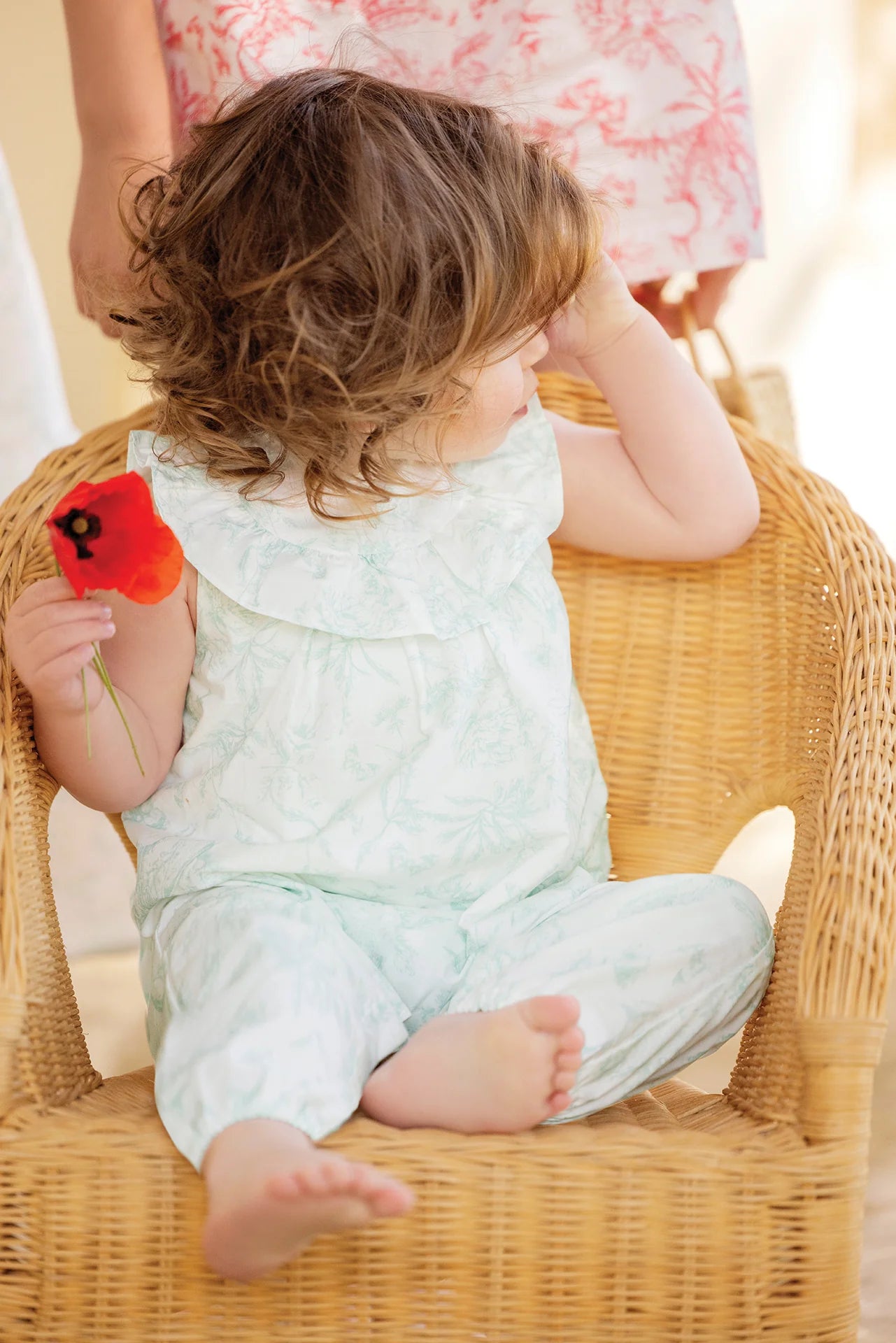 A young child with wavy brown hair sits in a wicker chair, wearing the Tartine et Chocolat Baby Girls' Sleeveless Jumpsuit and holding a red flower, looking to the side. An adult stands partially visible in the background.
