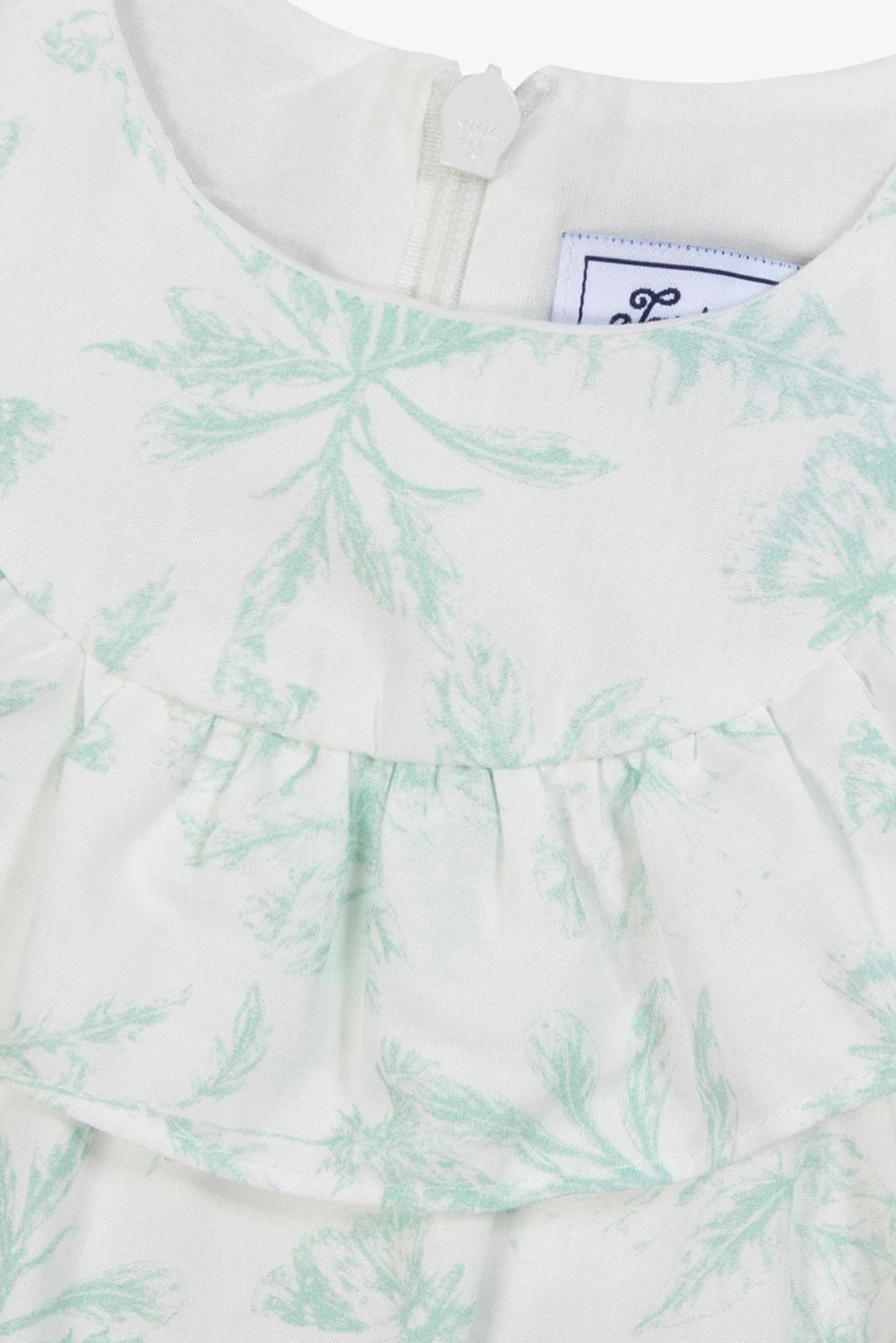 Close-up of the Tartine et Chocolat Baby Girls' Sleeveless Jumpsuit in white with a light green botanical Toile de Jouy pattern, ruffled detail, and back neckline zipper.