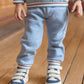 A boy wears the Tartine et Chocolat Baby Boys' and Boys' Jogger Outfit with a blue-grey sweatshirt and white sneakers with blue accents, standing on a wooden floor.