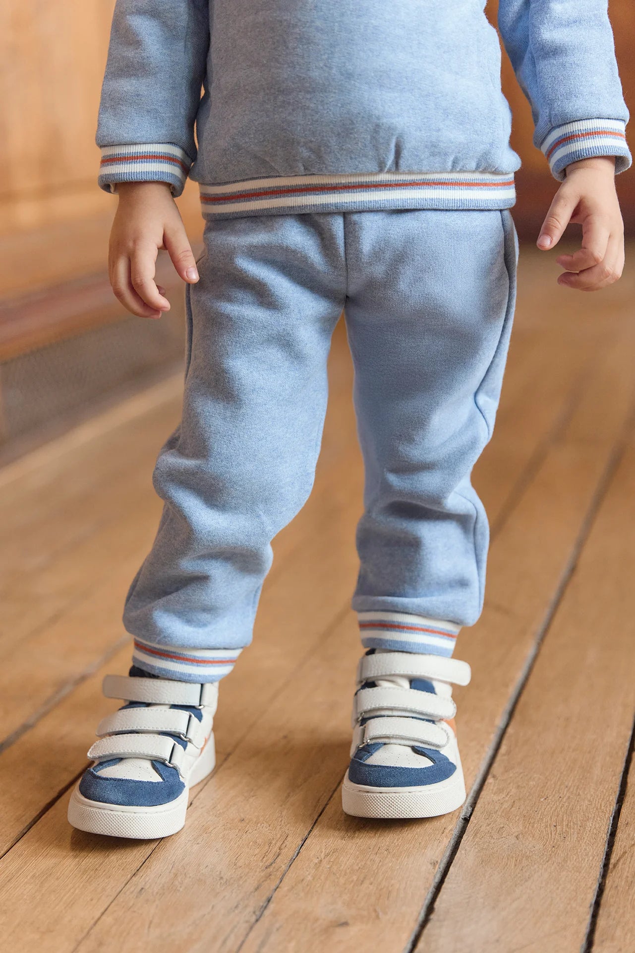 A boy wears the Tartine et Chocolat Baby Boys' and Boys' Jogger Outfit with a blue-grey sweatshirt and white sneakers with blue accents, standing on a wooden floor.