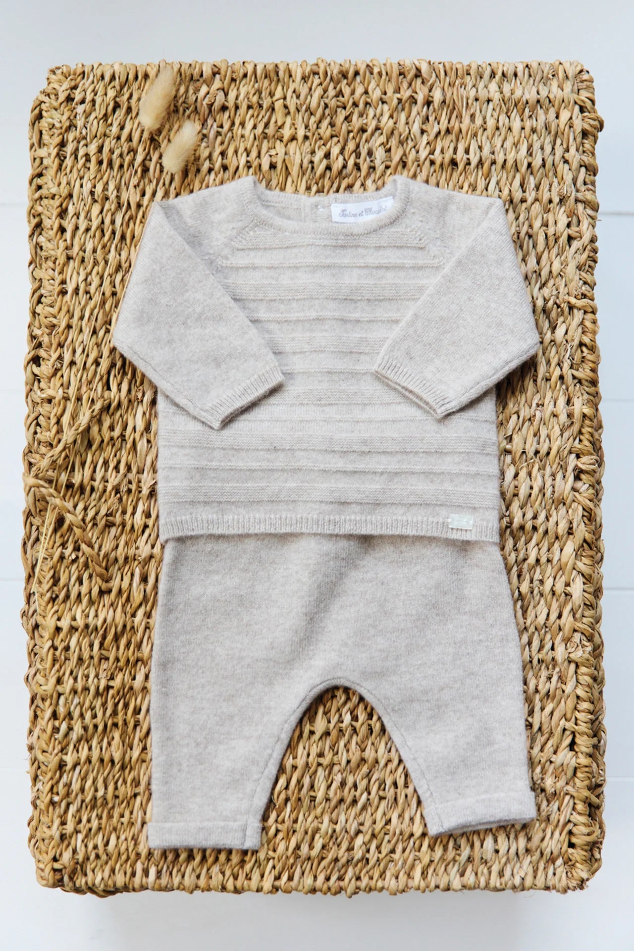 The Tartine et Chocolat Baby Long Sleeved Knit Outfit, featuring a beige sweater and matching pants in wool and cashmere, is displayed on a woven basket—a perfect winter choice for babies.