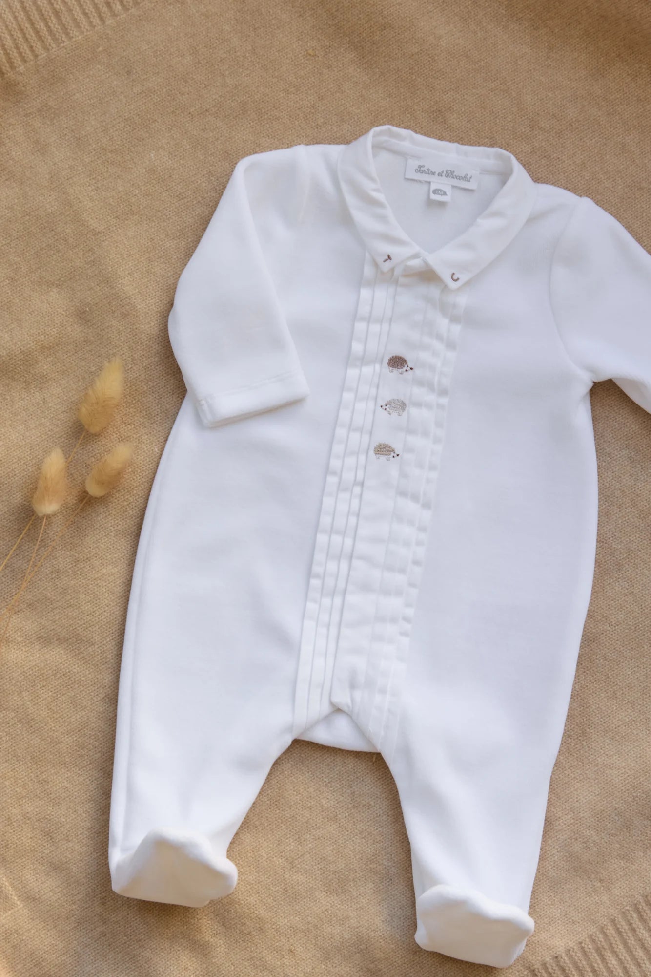 The Tartine et Chocolat Baby Boys' Hedgehog Pajamas—a cozy white long-sleeve onesie with pleated details and embroidered buttons—rests on beige textured fabric beside dried grass stems, perfect for snuggly nights.