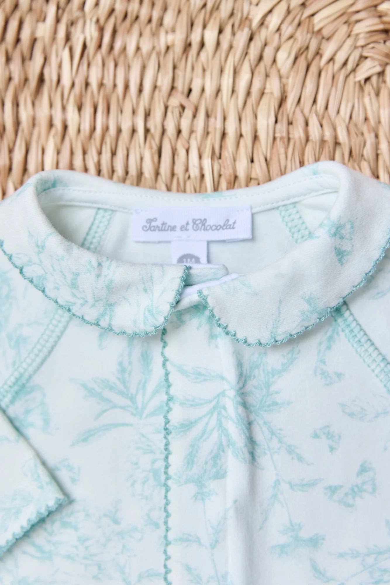 A close-up of Tartine et Chocolat Baby Girls’ Printed Pajamas, pale blue and white with a floral Toile de Jouy-inspired pattern and Peter Pan collar, displayed on a woven surface.