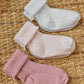 The Tartine et Chocolat Knit Sock 3-Pack features three pairs of soft cotton baby socks in white, pale pink, and mauve, neatly displayed on a woven wicker surface.