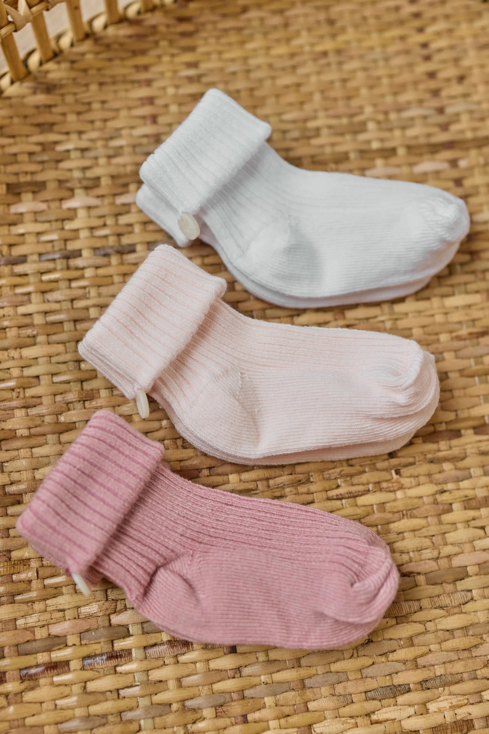 The Tartine et Chocolat Knit Sock 3-Pack features three pairs of soft cotton baby socks in white, pale pink, and mauve, neatly displayed on a woven wicker surface.