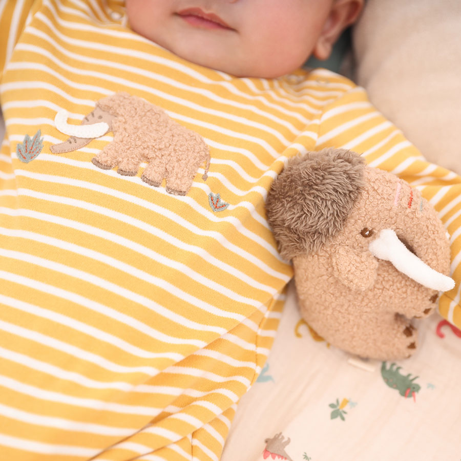 A baby is dressed in an Albetta Romper, Mammoth, featuring a charming yellow and white striped design made from premium BCI cotton with a delightful mammoth motif, lying beside a plush mammoth toy.