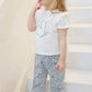 A young child with long blonde hair stands barefoot on white stairs, wearing the Tartine et Chocolat Baby Girls’ and Girls’ Liberty Butterfly T-shirt paired with patterned Liberty fabric pants, looking to the side with a finger near her mouth.