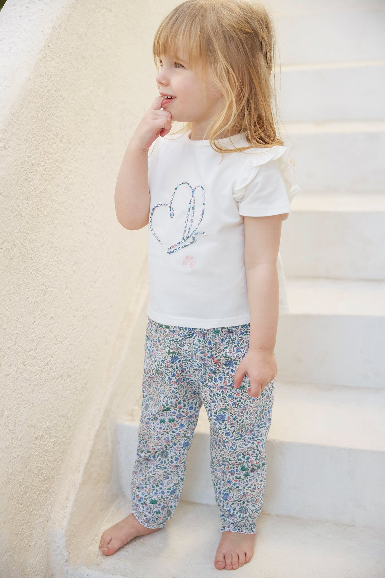 A young child with long blonde hair stands barefoot on white stairs, wearing the Tartine et Chocolat Baby Girls’ and Girls’ Liberty Butterfly T-shirt paired with patterned Liberty fabric pants, looking to the side with a finger near her mouth.