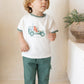 A young child with light brown hair stands barefoot indoors, wearing the Tartine et Chocolat Explorer Illustration T-shirt and green pants. A plant in a brown pot sits on a nearby ledge.