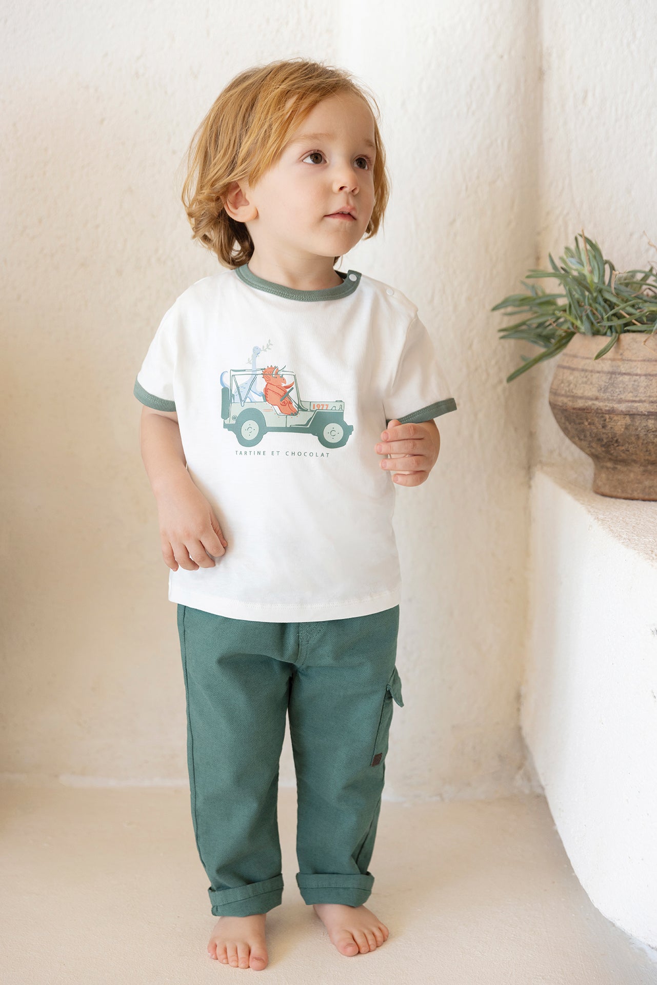 A young child with light brown hair stands barefoot indoors, wearing the Tartine et Chocolat Explorer Illustration T-shirt and green pants. A plant in a brown pot sits on a nearby ledge.