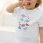 A young child wears the Tartine et Chocolat Baby Boys’ and Boys’ Illustrated Cotton T-Shirt, made from 100% cotton and featuring colorful dinosaur illustrations, paired with green shorts. Standing indoors with one finger on their lips, they’re ready for adventure—perfect attire for little explorers.