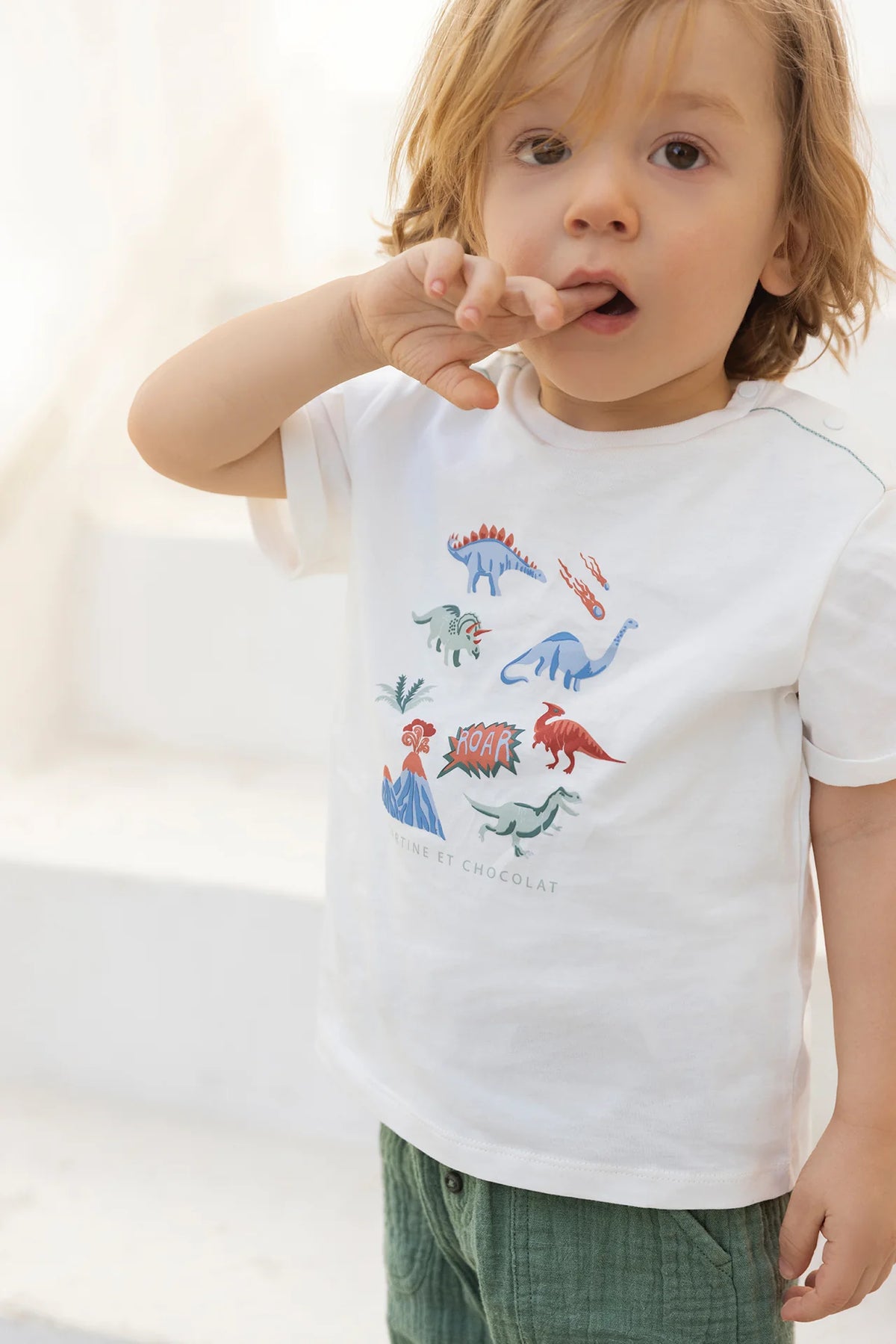 A young child wears the Tartine et Chocolat Baby Boys’ and Boys’ Illustrated Cotton T-Shirt, made from 100% cotton and featuring colorful dinosaur illustrations, paired with green shorts. Standing indoors with one finger on their lips, they’re ready for adventure—perfect attire for little explorers.