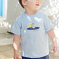 A young child with light brown hair stands outside wearing the Tartine et Chocolat Baby Boys’ and Boys’ Illustrated Cotton T-Shirt in light blue with a boat graphic, paired with navy shorts—an ideal outfit for little explorers. Sunlight and shadows create patterns on the wall and ground.