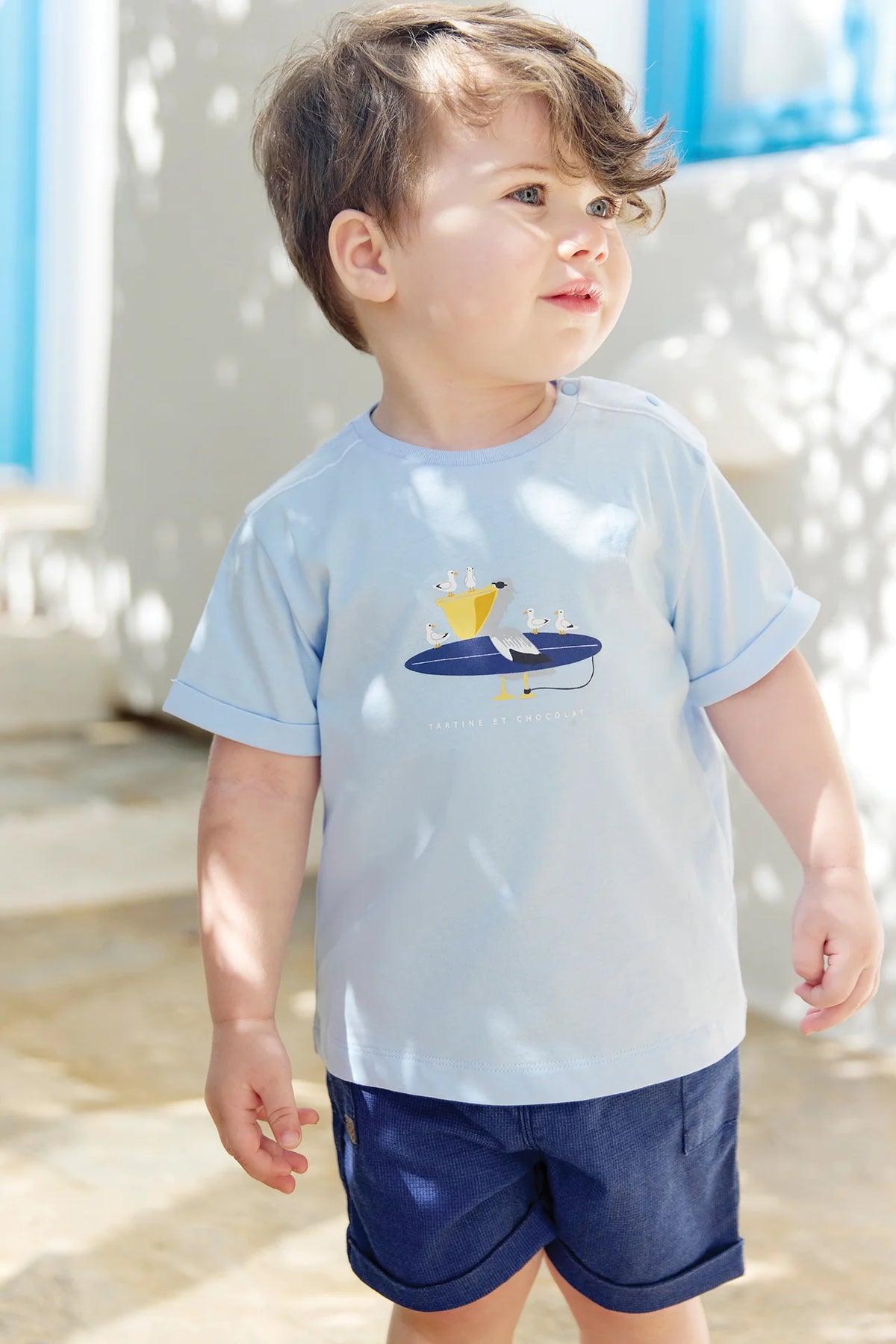A young child with light brown hair stands outside wearing the Tartine et Chocolat Baby Boys’ and Boys’ Illustrated Cotton T-Shirt in light blue with a boat graphic, paired with navy shorts—an ideal outfit for little explorers. Sunlight and shadows create patterns on the wall and ground.