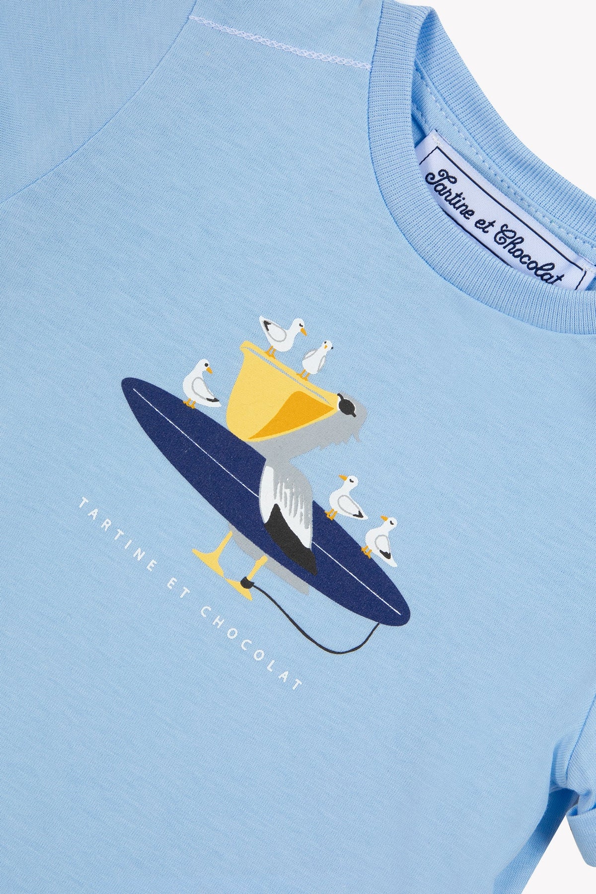 The Tartine et Chocolat Baby Boys’ and Boys’ Illustrated Cotton T-Shirt is a light blue top featuring a whimsical pelican riding a surfboard and carrying smaller birds in its beak, with “Tartine et Chocolat” printed below—a playful design made from 100% cotton, perfect for little explorers.
