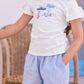 A young girl stands with one hand in her pocket, wearing a Tartine et Chocolat Girls’ Beach Illustration T-Shirt made from soft cotton and paired with light blue shorts; a wicker chair can be seen in the background.
