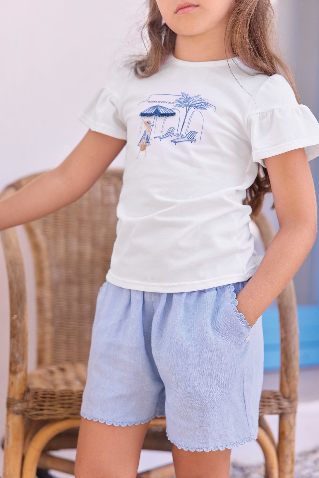 A young girl stands with one hand in her pocket, wearing a Tartine et Chocolat Girls’ Beach Illustration T-Shirt made from soft cotton and paired with light blue shorts; a wicker chair can be seen in the background.