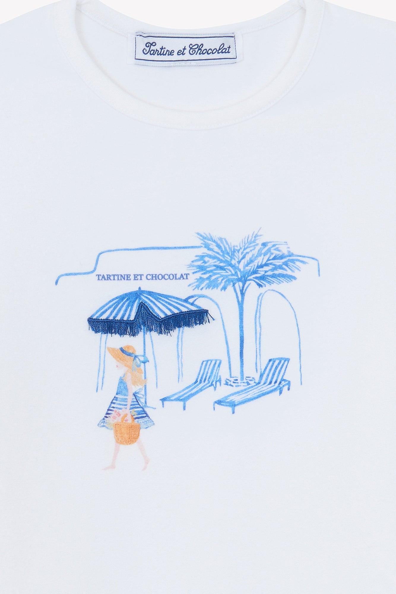 The Girls’ Beach Illustration T-Shirt by Tartine et Chocolat is a white cotton tee for girls, showcasing blue embroidery of a woman holding an umbrella, two lounge chairs, and a palm tree beneath the “Tartine et Chocolat” logo, with Santorini-inspired details for a charming seaside look.