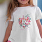 A young girl with long hair wears the Tartine et Chocolat Baby Girls’ and Girls’ Tropical T-shirt featuring colorful birds, an embroidered flamingo, and floral details. She pairs the summer cotton tee with pink pants and stands indoors against a white wall.