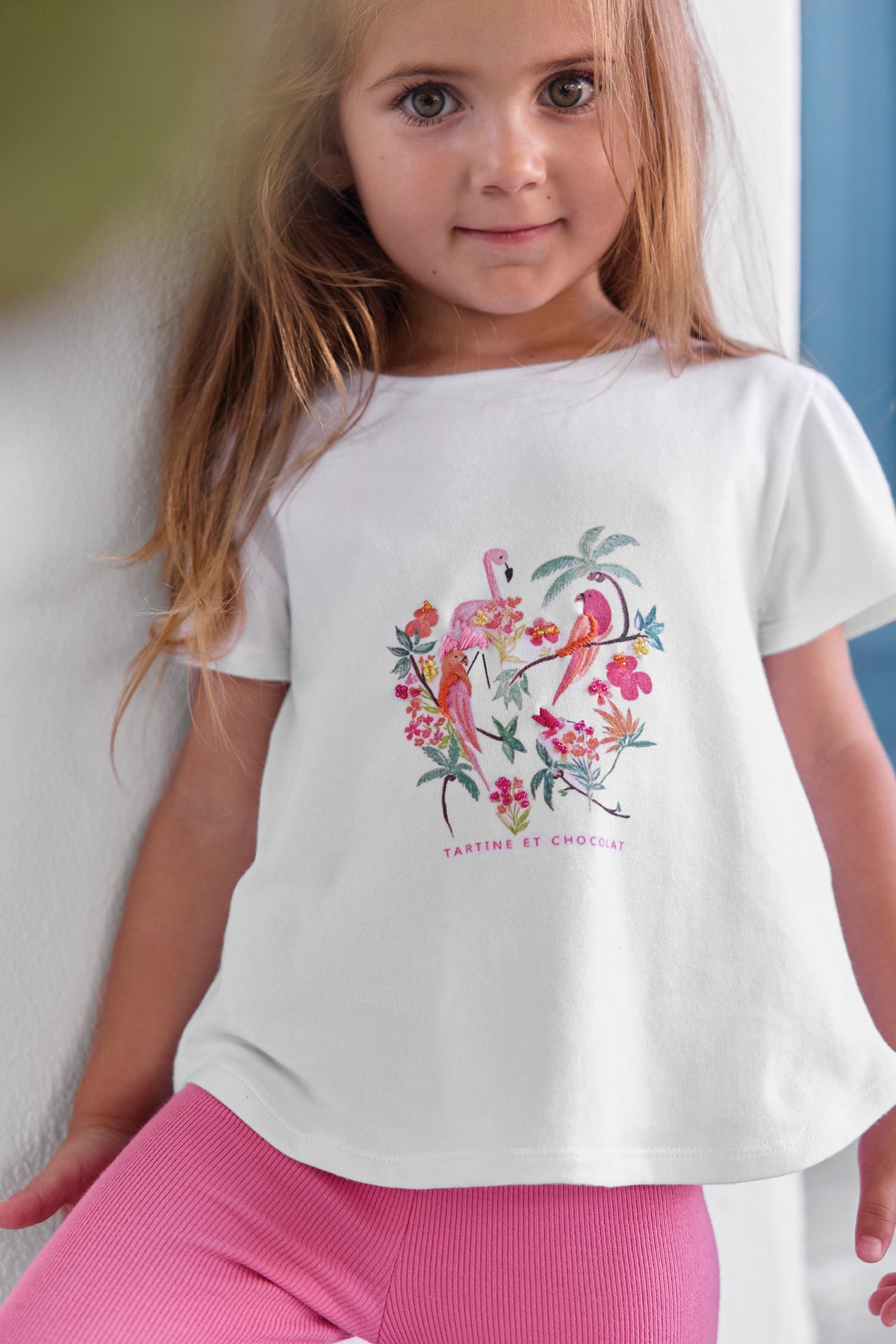 A young girl with long hair wears the Tartine et Chocolat Baby Girls’ and Girls’ Tropical T-shirt featuring colorful birds, an embroidered flamingo, and floral details. She pairs the summer cotton tee with pink pants and stands indoors against a white wall.
