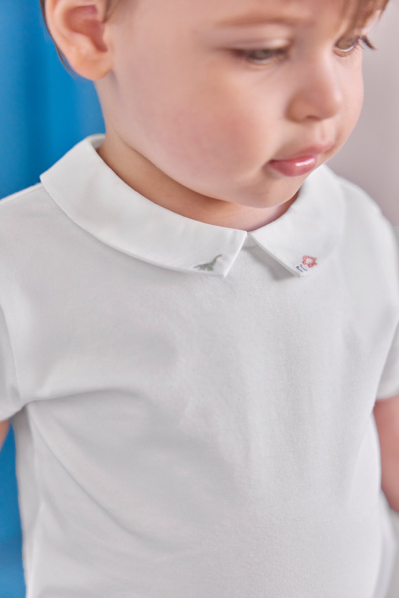 A toddler wearing the Tartine et Chocolat Baby Boys’ Pointed Collar Onesie in ecru with a delicately embroidered collar, gazing downward.