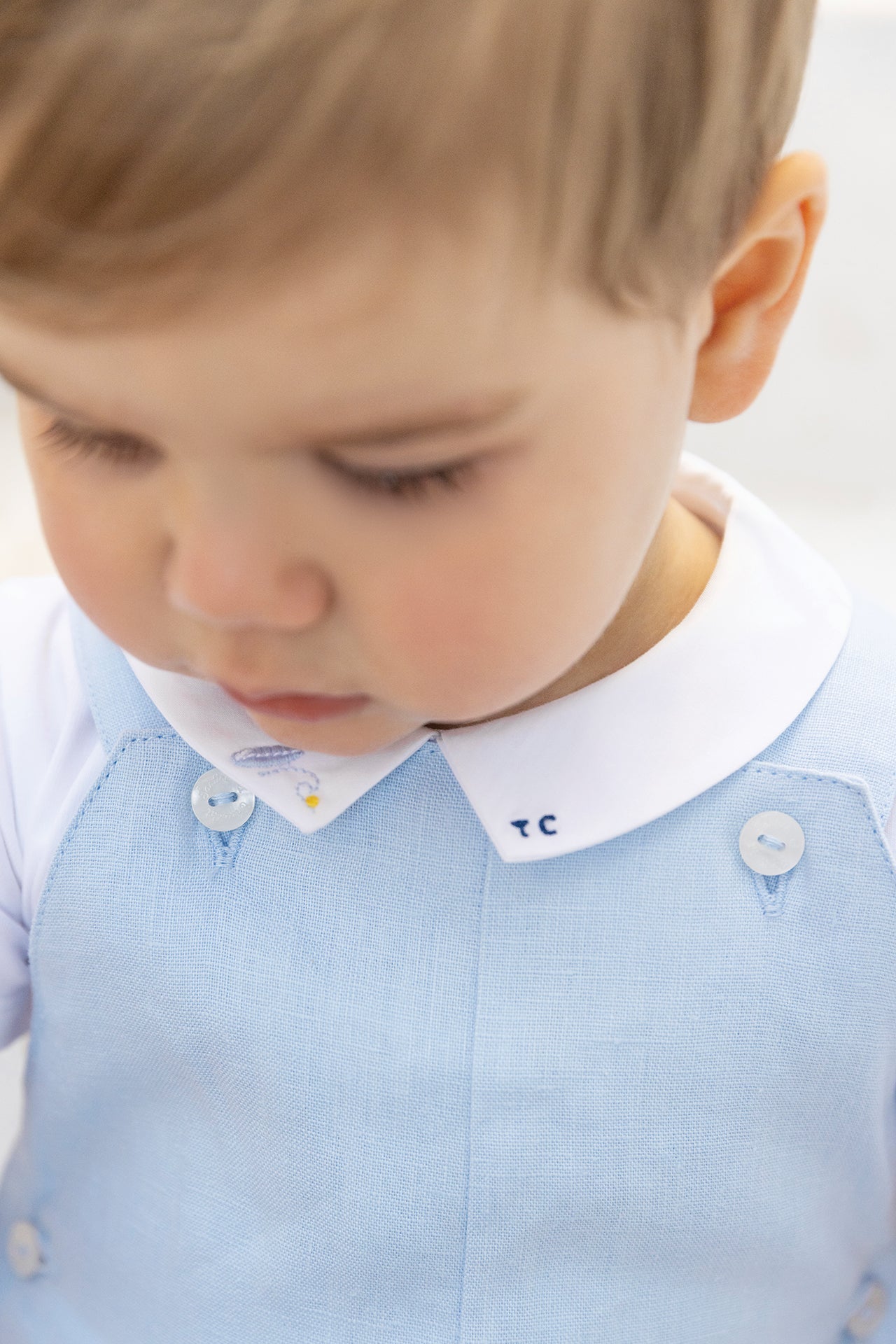 A close-up of a toddler wearing the Tartine et Chocolat Baby Boys’ Pointed Collar Onesie, featuring a light blue design with white buttons and an embroidered collar, looking downward.