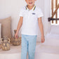 A young boy with short brown hair stands barefoot on white steps indoors near decorative pottery, wearing the Tartine et Chocolat Boys’ Surfer Polo Shirt with a striped collar and light blue pants.