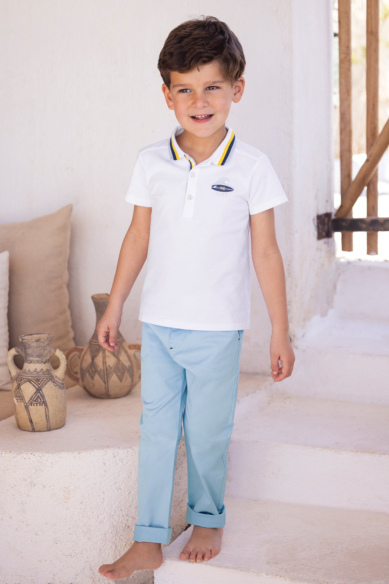 A young boy with short brown hair stands barefoot on white steps indoors near decorative pottery, wearing the Tartine et Chocolat Boys’ Surfer Polo Shirt with a striped collar and light blue pants.