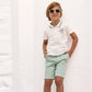 A young boy with sunglasses, dressed in a white polo shirt and light green Boys’ Cotton Short from Tartine et Chocolat, stands barefoot against a white wall with his hands in his pockets, smiling. The breathable fabric of the shorts ensures comfort for active play all day long.